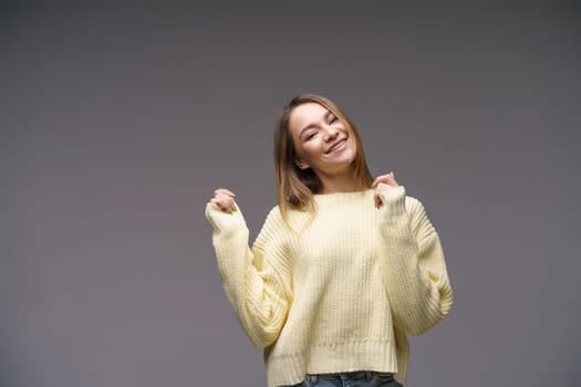 Portrait of a beautiful young girl of Caucasian ethnicity in a yellow sweater on a gray background cute smiling. Keeps his hands to his face.