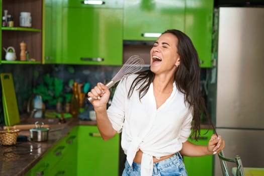 Happy young woman holding whisk as microphone singing song, dancing, cooking alone in modern kitchen funny housewife lady having fun listening to music, preparing healthy morning meal, doing housework