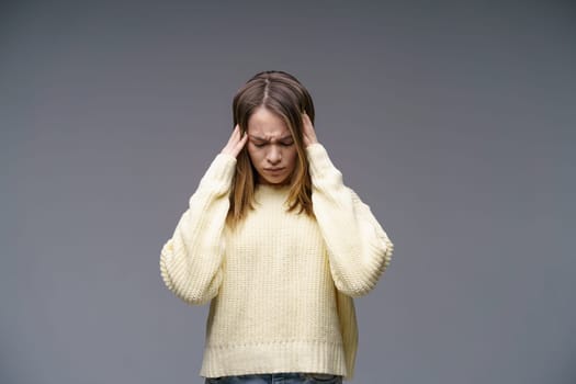 A young woman of Caucasian ethnicity is holding her head and squeezing her eyes in pain in a yellow sweater on a gray background,