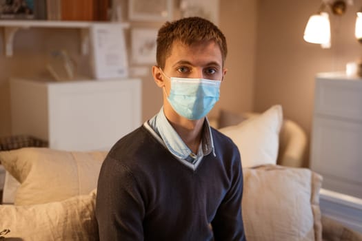 a young guy of Caucasian ethnicity in a shirt and a protective mask sits on the couch alone at home. Concept of being at home during global quarantine