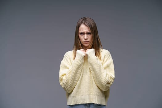 A young woman of Caucasian ethnicity, very upset in a yellow sweater on a gray background, holds her hands in fists. Emotional experiences and problem solving