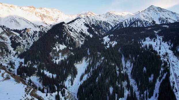 Coniferous forest in snowy mountains. White clouds float across the sky. Top view from a drone. In places, the road to the mountains and buildings are visible. There is a pipe along the gorge. Sunset