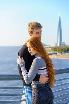 A guy and a girl of Caucasian ethnicity stand together on a river bridge on a sunny day with a city view