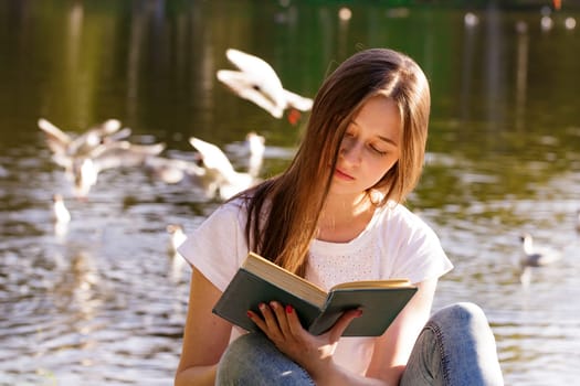 Young caucasian woman sitting on the shore of the lake reads a book on a sunny day. Reads outdoors in the park