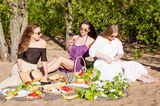 Friends having fun at beach on sunny day. Party time. Summer holidays vacation happy people concept. Three young female friends is having picnic with wine, cheese and fresh fruits on hill at sunset.