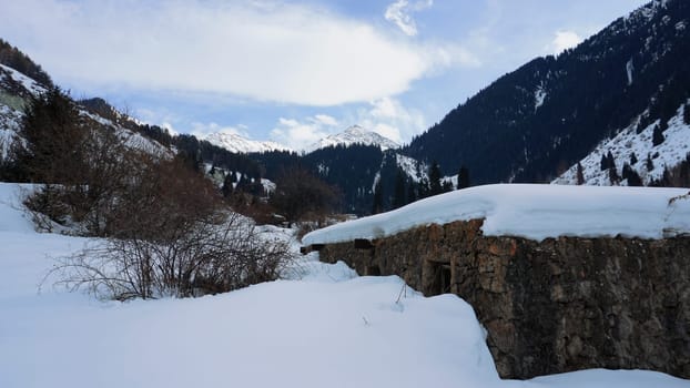 An old stone prison or hut in the mountains. Snowy mountains, forest and clouds against a blue sky. Tall dry bushes peek out from under the snow. Birch and spruce trees are visible in the distance.