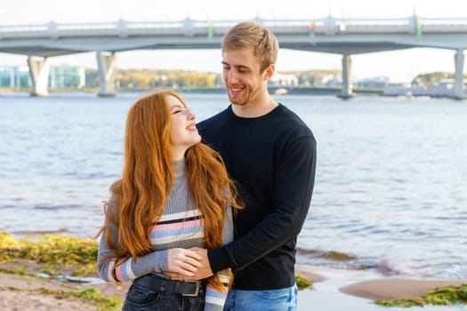 Young couple of man and woman with long red hair of Caucasian ethnicity, in casual clothes, stand on the bank of the river embracing happy on a summer day against the background of the cityscape