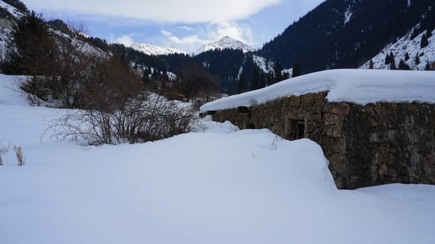 An old stone prison or hut in the mountains. Snowy mountains, forest and clouds against a blue sky. Tall dry bushes peek out from under the snow. Birch and spruce trees are visible in the distance.