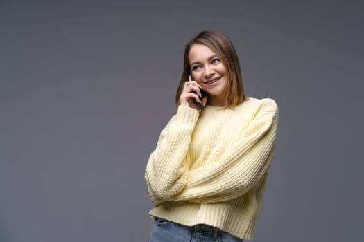 Young beautiful woman of Caucasian ethnicity talking on the phone in a yellow volumetric sweater on a gray background with blue eyes