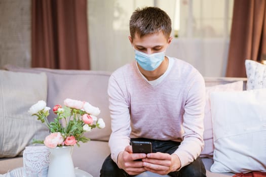 young caucasian man sitting on a sofa in casual clothes, wearing a protective medical mask with a phone in his hand