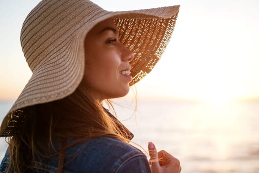 Portrait of a happy elegant young woman in a white denim jacket and a straw hat on the ocean shore at sunset, looking into the distance. young caucasian female model close up on the seashore.
