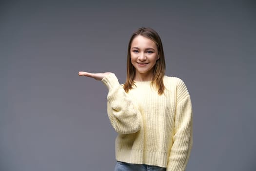 Portrait of a young woman of Caucasian ethnicity shows with her hand to the side in a yellow sweater on a gray background, the concept of advertising goods on her hand