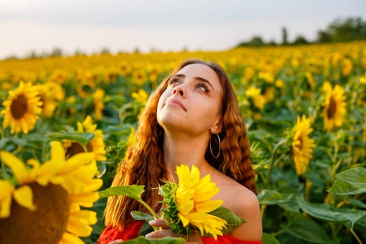 Female portrait of young woman in field of blooming sunflowers in rays of sunset . Cute girl of Caucasian appearance in evening, free and happy in red dress, stands in a field of bright sunflowers