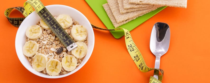 oatmeal with sliced banana and crispbread lie with spoon and yellow measuring tape on orange background, healthy food and diet concept