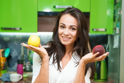 Young woman stands in kitchen holding fruits in her hands. Caucasian girl in a modern kitchen holds green apple and ripe peach in her hands. Chooses what to eat. Healthy nutrition for beautiful skin