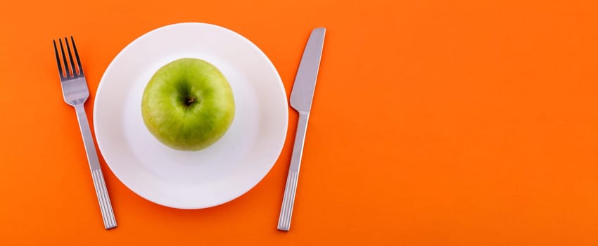 On a white plate lies a green apple, a knife with a fork on an orange background, view from the top. Diet and healthy food concept