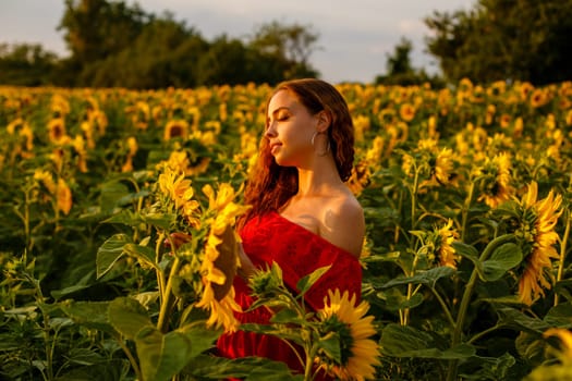 Young woman smiling at sunset in a field of sunflowers. A cute girl of Caucasian appearance in a red dress is happy and free to stand in the evening in the rays. Natural beauty in nature
