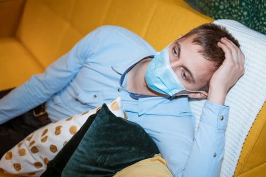 a young guy of European appearance, sitting comfortably on the couch in a medical mask. Concept to wait out the pandemic while sitting at home