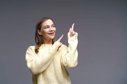 Cheerful young woman of Caucasian ethnicity shows thumbs up in a yellow sweater on a gray background, positive advertising. Beautiful girl posing in studio