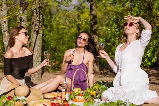 Cheerful girlfriends have fun in summer at picnic. Cheerful company of beautiful girls. Young caucasian women wearing sunglasses and drinking wine, drinking wine and eating fresh fruits in nature.
