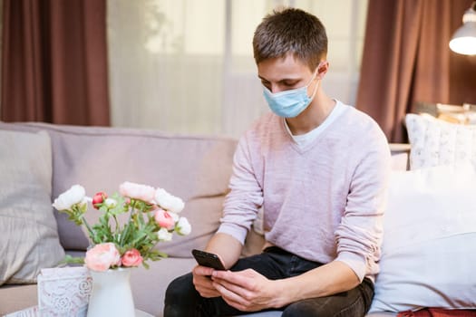 young caucasian man sitting on a sofa in casual clothes, wearing a protective medical mask with a phone in his hand