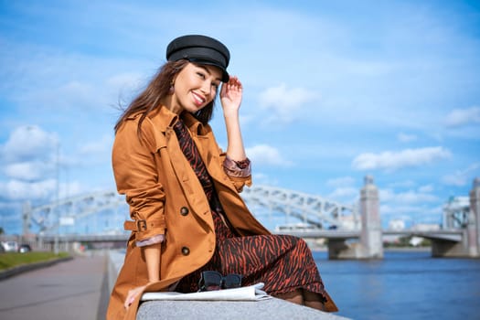 portrait of a happy Caucasian woman on the embankment of the river in a black cap and brown jacket on a sunny day