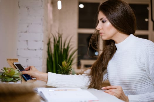 Beautiful business woman in a white sweater is working on the online communication by phone while sitting at the table