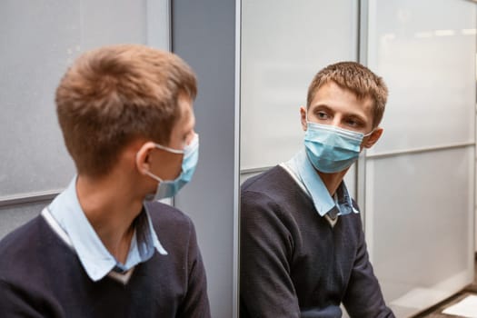 young caucasian man in a protective mask looks in the mirror, adjusts the medical mask against the virus. The concept of wave ipidemii