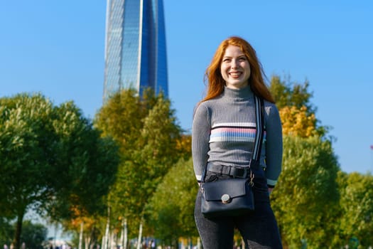 Redhead happy woman of Caucasian ethnicity with bag posing in park on green grass on sunny spring day. Cute girl in the city park against the background of green trees and blue sky