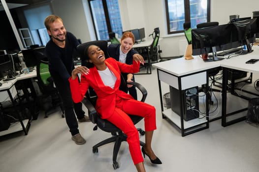 Caucasian red-haired woman, bearded caucasian man rolled African American young woman on office chair. Colleagues have fun at work