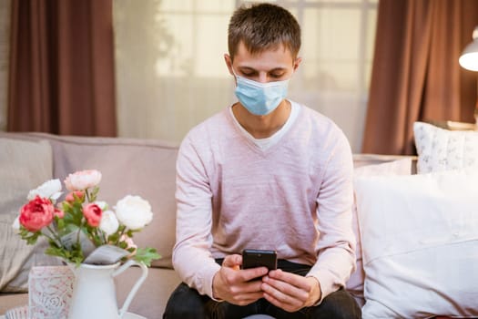 young man of European appearance in casual clothes, at home on the couch in a medical mask communicates by video communication on the phone, the concept of remote online communication on the Internet