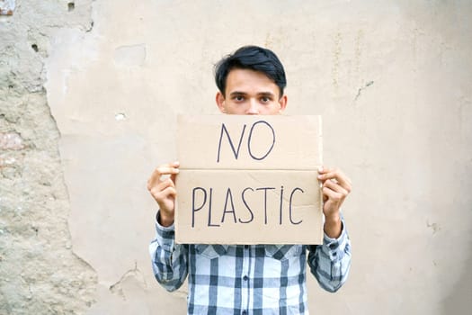 Caucasian guy holding the inscription no plastic. Environmental protection concept from garbage pollution . Young man protesting with placard banning plastic against cement wall background