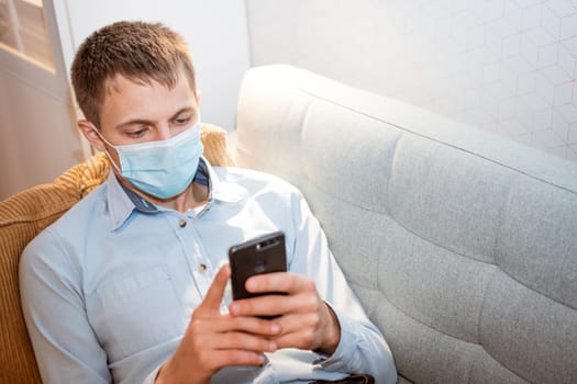 young caucasian man sitting on a sofa in casual clothes, wearing a protective medical mask with a phone in his hand