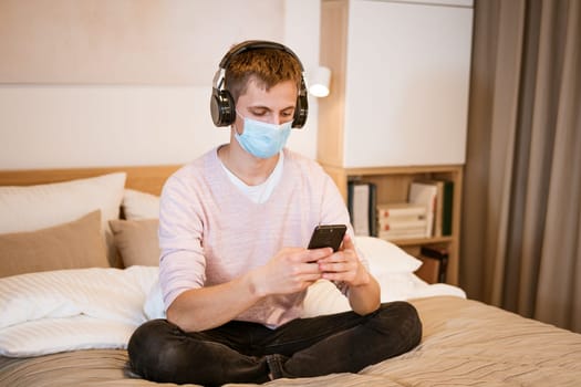 a guy of Caucasian nationality in casual clothes, wearing headphones on the bed in a protective medical mask with a phone in his hand, watching a video online on a Wi-Fi network