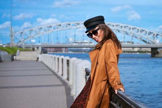 Beautiful caucasian woman in sunglasses, in a black cap and jacket posing while standing on the embankment on a sunny day against a background of blue sky and cityscape