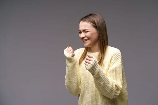 A young woman of Caucasian ethnicity, very upset in a yellow sweater on a gray background, holds her hands in fists. Emotional experiences and problem solving