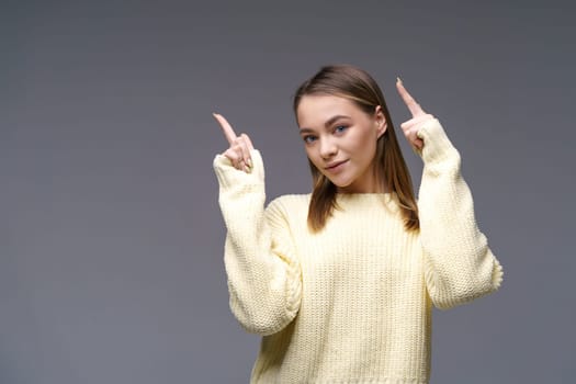 Cheerful young woman of Caucasian ethnicity shows thumbs up in a yellow sweater on a gray background, positive advertising. Beautiful girl posing in studio