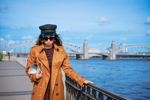 Young beautiful caucasian woman in a black cap and sunglasses in a coat on the embankment posing with a newspaper in her hand against the background of a blue sky and a bridge