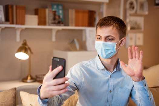 young man of European appearance in casual clothes, at home on the couch in a medical mask communicates by video communication on the phone, the concept of remote online communication on the Internet