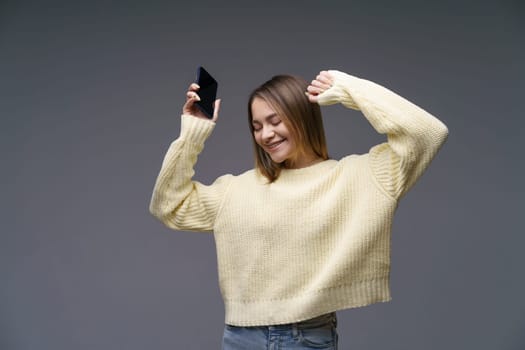 Young cheerful woman of Caucasian ethnicity in a yellow sweater on a gray background is dancing with a phone in her hand. Happy girl listening to music on the phone dancing raising her hands up