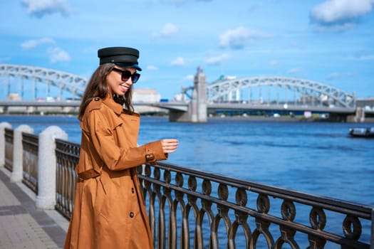 Happy young woman of Caucasian ethnicity in a light brown coat and black cap posing on the embankment against the backdrop of the bridge and the blue sky.
