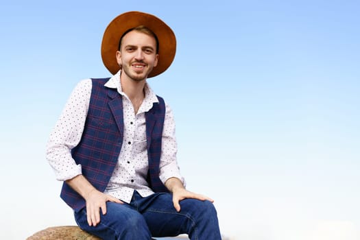 Portrait of a handsome young man in a hat against a blue sky. Caucasian guy with a goatee smiling happy summer outdoors sitting in a shirt and vest