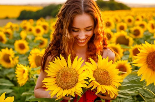 Cute young woman is holding sunflower in her hand while standing in field at sunset. Beautiful gentle girl of Caucasian ethnicity in red dress in the rays of setting sun. The concept of natural