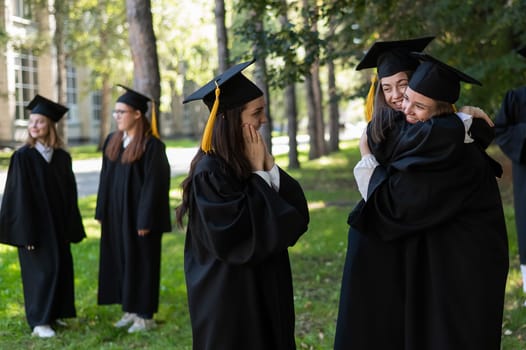 Group of happy students in graduation gowns outdoors. In the foreground, two young girls congratulate each other and embrace