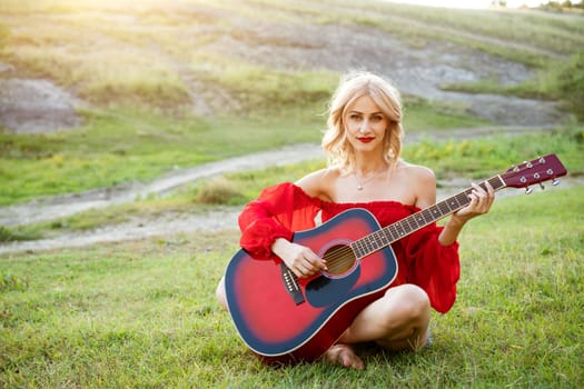 The woman in red is sitting on the green grass with a red guitar. Girl musician plays the guitar outdoors. Blonde caucasian appearance