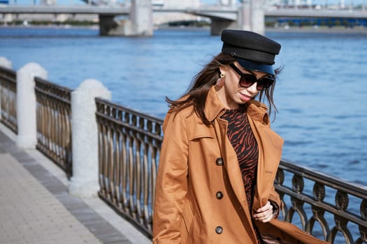 A female portrait of European appearance on the embankment on a sunny spring day with a newspaper in her hand. Beautiful brunette woman in sunglasses and in a brown jacket and black cap