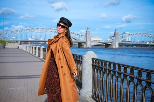 Happy young woman of Caucasian ethnicity in a light brown coat and black cap posing on the embankment against the backdrop of the bridge and the blue sky.