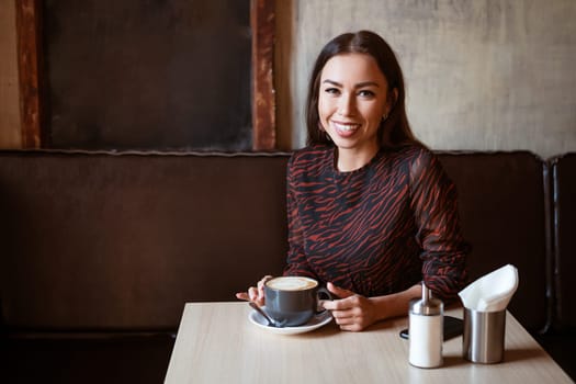 A young beautiful woman with dark hair, Caucasian appearance with makeup in a brown dress is sitting at a table in a cafe with coffee and smiling with a snow-white toothy smile