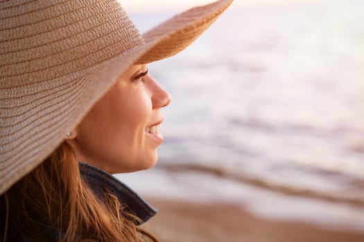 Portrait of a happy elegant young woman in a white denim jacket and a straw hat on the ocean shore at sunset, looking into the distance. Young caucasian female model close up on the seashore.