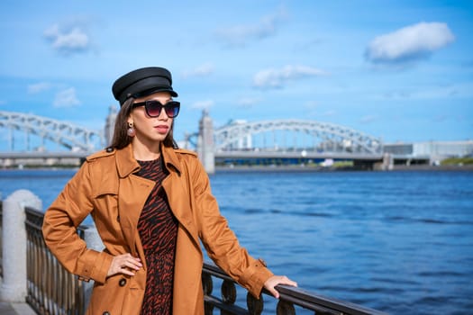 A young European woman in sunglasses and a black cap poses on the embankment in a brown coat on a spring sunny day. The concept of a successful and free woman
